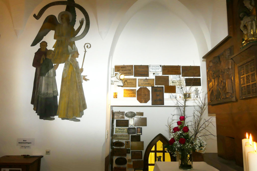 Innenansicht einer Kirche mit Gemälden, Votivtafeln und roten Rosen.