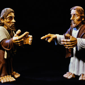 Zwei lebensgroße, barttragende Figuren in Umhängen gestikulieren vor schwarzem Hintergrund.