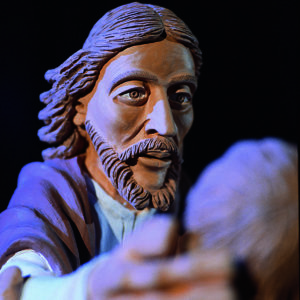 Eine Skulptur Christi vor Schwarz zeigt expressiven Blick und ausgestreckte Hand.