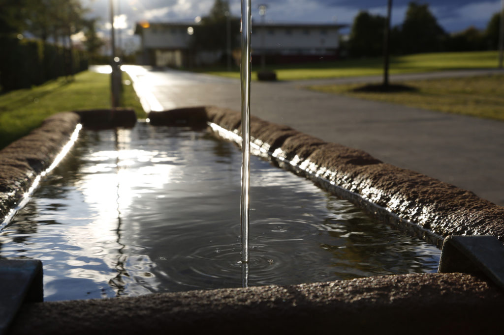 Ein steiniger Wasserlauf mit zentralem Wasserstrahl steht im Vordergrund vor einem Gebäude.