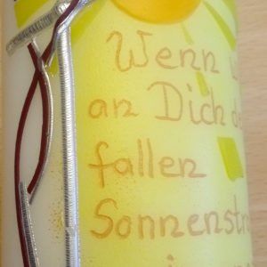 Eine Kerze mit Sonnenmotiv und Spruch hängt vor hellem Hintergrund.
