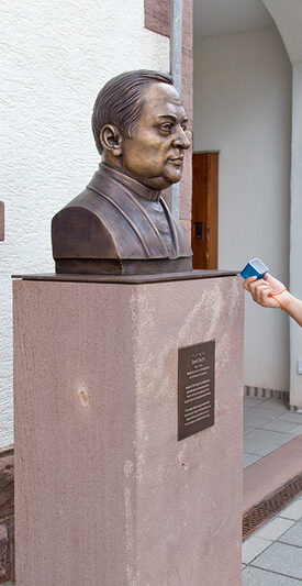 Eine Frau mit blauem Gerät an einen bronzenen Kopf am historischen Rundweg vor einem Backsteinbau.
